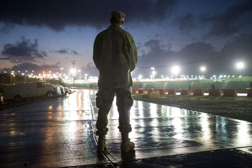 A soldier, back to the camera, stands amid the glare of lights reflecting off the wet concrete.