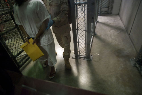 A shackled detainee, holding a manila envelope, is ushered through a gate.