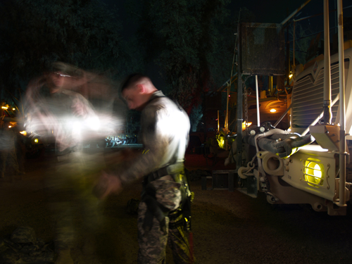 American soldiers gear up by the headlights of their armored vehicles.