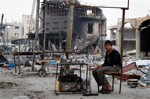 A sandal-wearing boy sits on a crude, battered chair, a small, equally-battered table before him. Behind him is the rubble of large, urban structures.