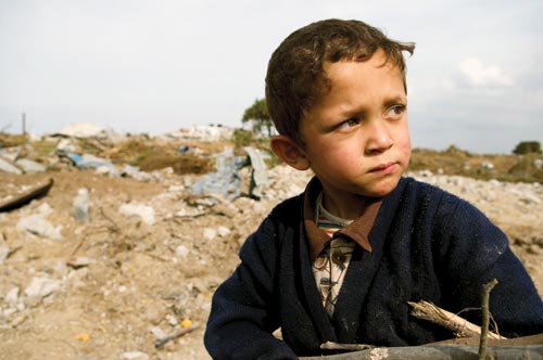 A young boy, not much older than a toddler, holds a bundle of firewood against his chest. Behind him is debris. He wears a sweater over a button-down shirt and, beneath that, a rainbow-striped undershirt.