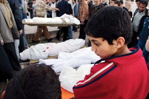 A boy stands in front of several bodies, each wrapped in white cloth. The corpses are surrounded by men, all looking downward. The boy averts his gaze.