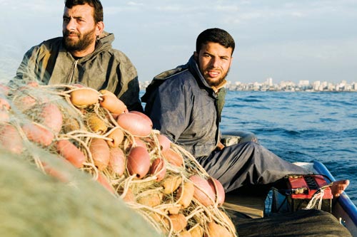 Two bearded men sit at at the stern of a boat. In front of them is a large bundle of netting and buoys.