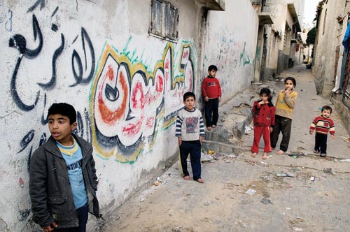 A group of young children congregate in an alley. Several of them are barefoot. The walls of the buildings are graffitied.