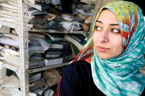 A young woman, wearing a headscarf, stands in front of a shelf weighed down with ragged books.