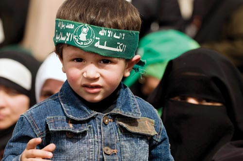 A young boy wears a green headband. Behind him are hijab-wearing women.
