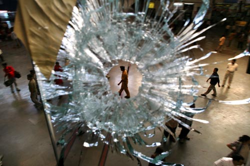 An officer patrols a train station, as seen through a bullet hole in glass above.