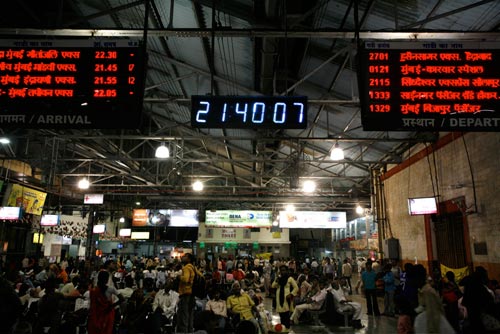 A train station is crowded with people. Arrival and departure boards loom over them, suspended from a corregated metal roof.