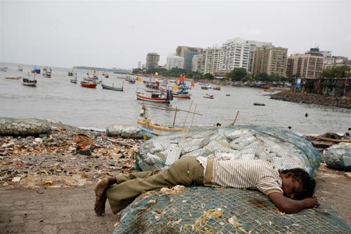 At the water's edge, a young man sleeps on a fishing net. Behind him is a filthy shore, and then a small inlet crowded with small boats. On the other side of the inlet is a cluster of squat high-rise buildings.