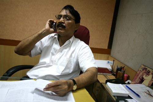 A middle-aged man, sitting at a desk, talks on a mobile phone. The top of his desk consists of a single pane of glass. Papers are spread out on it. Off to one side is a photograph of a medal being pinned onto his chest; he's wearing a khaki police uniform.