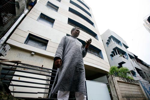 Standing next to a six-story building, a man looks at his mobile phone. He's wearing a kurta, a knee-length shirt not unlike a nightgown.
