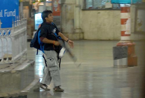 A young man walks through a train station. Nobody else can be seen. He's carrying a large backpack. In his right hand is a large rifle with a sizable clip of ammunition.