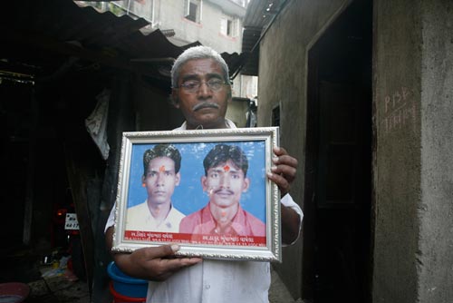 A middle-aged man stands outside of a ramshackle garage, holding a large, framed portrait of a pair of young men. He looks sad.