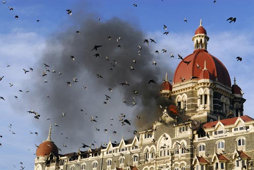 An ornate, castle-like hotel is on fire, a cloud of black smoke rising to meet the blue sky. In the foreground, a flock of birds flies by.