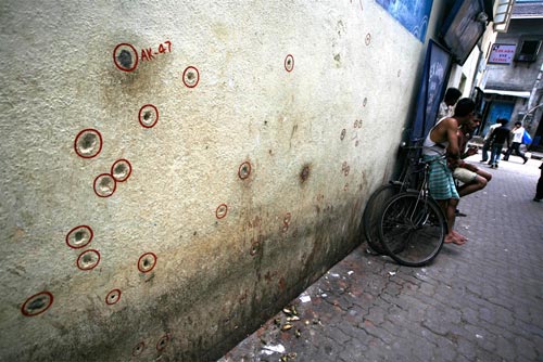 Men stand in an alleyway, and people can be seen walking by on a perpendicular street. The wall is cratered with dozens of bullet holes, sprayed over a fifteen-foot-wide, six-foot tall segment. Each of them is circled in red, and one of them is labelled 'AK-47'.