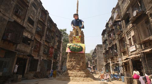 In a wide alley between rows of shabby four-story residential buildings, a three-story-tall hay bale pile supports an effigy of the Mumbai terrorist caught on camera, wearing his distinctive blue shirt, carry a backpack, and holding a silver gun. Below him is a large sculpture of a grenade.