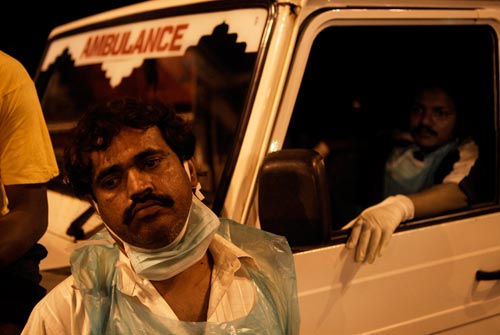 A dejected-looking man stands in front of an ambulance. A surgical mask is looped around his ears, but it's pushed down under his chin. He wears a disposable plastic smock.