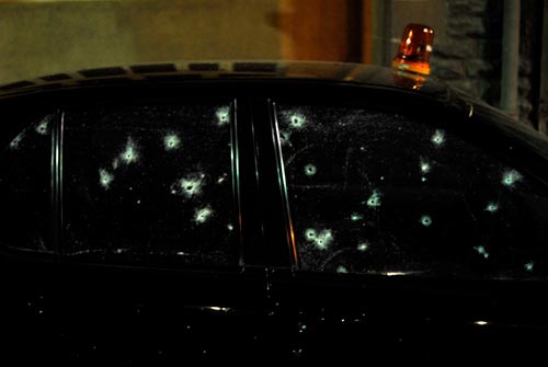 A black police car with darkly tinted windows is riddled with bullet holes. A red siren, intact, perches atop the roof.