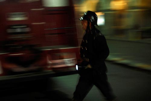 A man dressed in the manner of an English Bobby, carrying a flashlight, walks past a fire truck. His mouth is open, as if shouting.