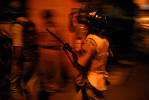 A trio of soldiers rush through the night, wearing bulletproof vests and helmets, carrying rifles.