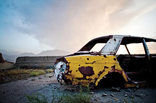 The rusted yellow-and-white body of a car, all other parts gone, rests on concrete. In the background is a mountain range, and the beginnings of a sunset.