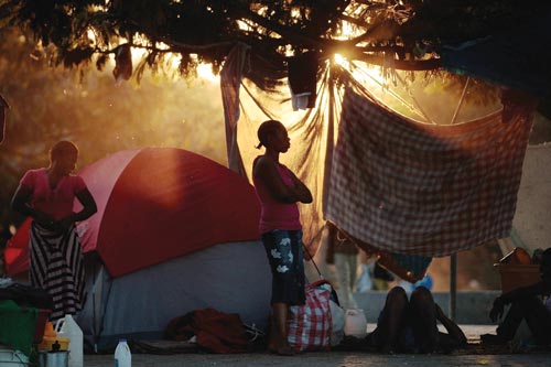 Women stand in the orange, dying light in front of a tent. Jugs and bags are on the ground around them. In the background are more people and more tents.