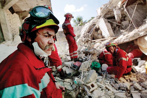 A half dozen red-suited, helmeted men surround a small opening in a pile of rubble.