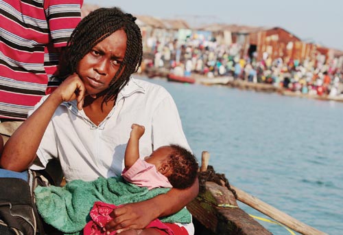 A serious-looking woman sits on a boat, head resting on her hand, holding a baby on her lap. The shore is only a few hundred yards away.