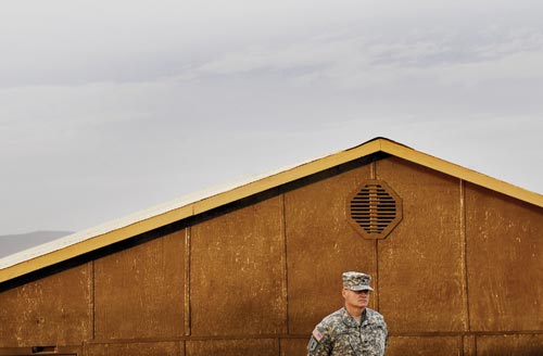A uniformed American soldier stands at attention in front of a low building.