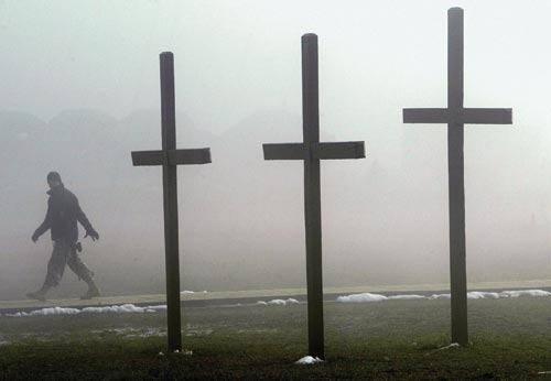 Three simple crosses, perhaps ten feet tall, are planted in the grass. Remnants of snow are near their base. In the background, a soldier walks by, obscured by fog.