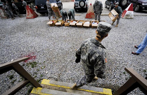 Shipping boxes are lined up in a gravel yard, where several soldiers look through the contents.