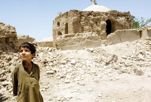 A young boy, wearing a brown galabiyya, stands in front of a pile of rock rubble, behind which are the badly-damaged remains of a simple mosque.