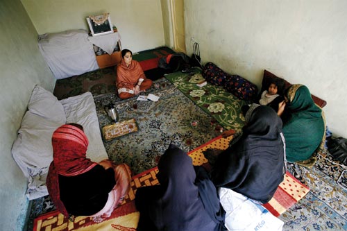 A woman sits crosslegged on the floor of a sparsely-decorated room. Across from her are four women and a child, facing her.