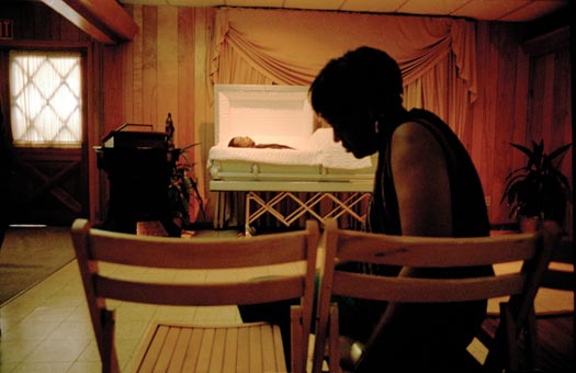 In the background, half of a casket is open, displaying the body within. In the foreground, a black-clad woman sits on a folding chair. The room has a tile floor, light-colored wood walls, and a drop ceiling.