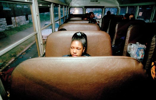 A girl sits alone on the brown pleather seat of a school bus, looking out the window. She's one of just a handful of students on the bus. It is twilight outside.