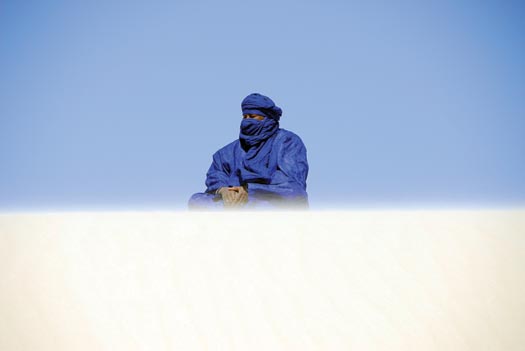 A person in cobalt-blue robes sits cross-legged on the sand.