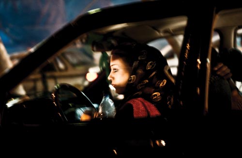A young woman sits behind the wheel of a darkened car. Her headscarf has slipped back, uncovering most of her head. She holds a bottle of water in one hand. A passenger is in the car as well.
