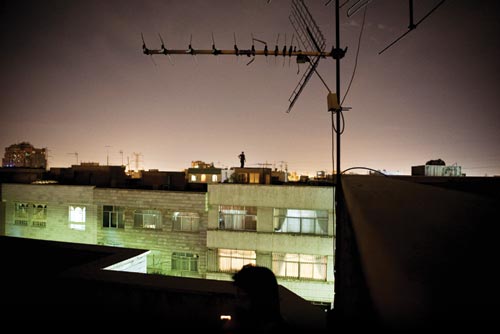 From one rooftop, the rooftop of an adjacent building can be seen. Silhouetted against the glow of city lights at night is the figure of a lone man, one hand on his hip.