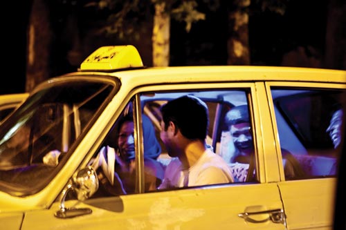 A yellow taxi is full of laughing young adults. A young man drives, a smiling young woman sitting next to him.