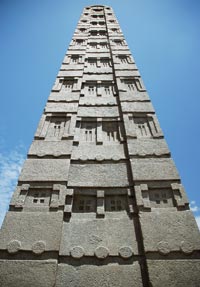 Looking straight up the face of the obelisk as it rises into the blue sky. Up close it can be seen that the scalloped pattern comes from shadows cast by button-like discs that emerge in relief from the surface.