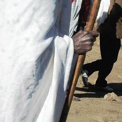 A dark-skinned old man leans on a cane as he walks. Nearly all of his features are out of the frame: we see only his enveloping white garment and one wrinkled hand. In the background are the lower bodies of two other people walking across the sun-drenched dirt.