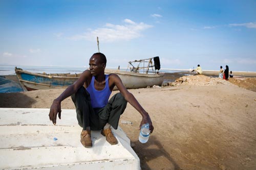 A young man squats on an overturned boat, a bottle of water in his hand. He's wearing a blue sleeveless t-shirt and a worn-looking pair of dress shoes. Behind him can be seen a few other small boats and a few people looking out at the sea.