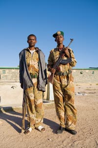 Two young soldiers pose for the camera. Both are wearing oversized desert-style camouflage. One has a green beret, an automatic weapon with a banana clip, and black dress shoes. The other is hatless, wears a gray windbreaker and white sandals, and carries an ancient-looking rifle.