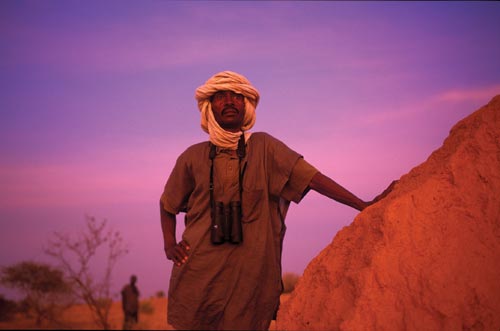 A moustached man leans against a rock, looking into the distance. His head is wrapped in a white scarf. A pair of binoculars hang around his neck. The sun, presumably low on the horizon, gives everything a reddish cast.
