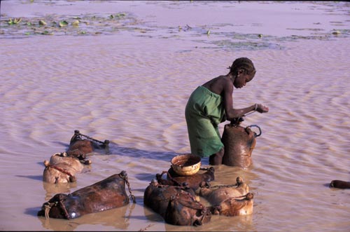 Standing in shin-high muddy water, a girl is surrounded by bags that she's filling with water. Each of the bags is othing more than the skin from the body of an animal, the legs tied off with rope.