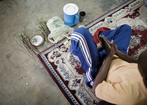 A young man sits on a woven rug, hands clapsed together as if in prayer. Laid out before him are a pack of cigarettes, a bunch of dried plants, a pitcher, and a cup.