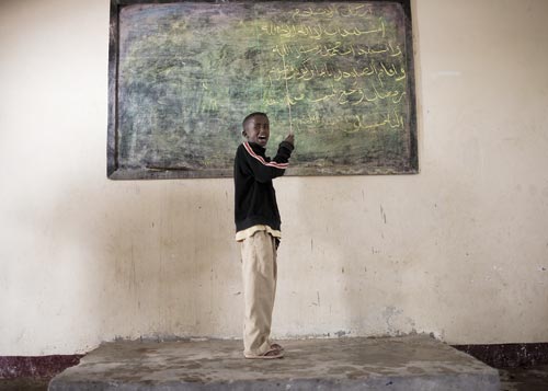 A boy, clad in khakis and a sweater, stands in front of a chalkboard, chalk in hand.