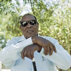 A well-fed, middle-aged man wears sunglasses and leans on a cane. He wears a white button-down shirt and sits in shade under a tree.