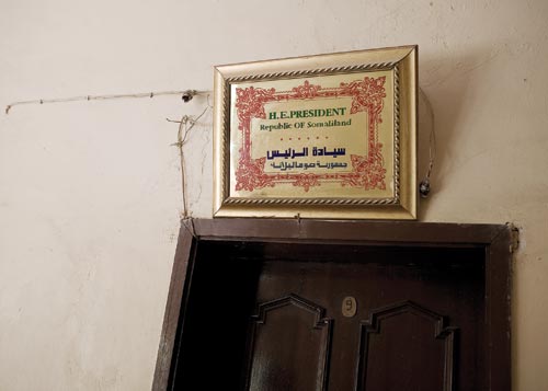 A certificate hangs on a cracked wall above a door, surrounded by exposed wiring. It reads “H.E.PRESIDENT,” and “Republic OF Samalialand.”