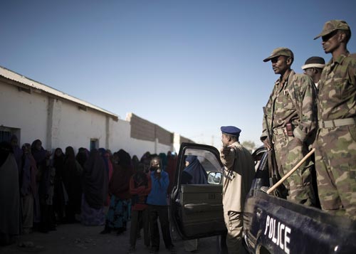 Camouflage-uniformed police officers stand in the bed of a pickup truck, carrying sticks and rifles. A long line of women stands in the shade of a building. One of them talks to a gesticulating officer.